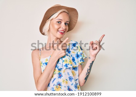 Young blonde woman with tattoo wearing summer hat smiling and looking at the camera pointing with two hands and fingers to the side. 