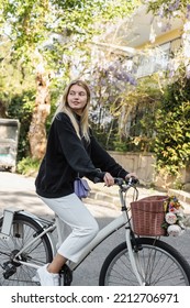 Young Blonde Woman In Trendy Outfit Riding Bicycle Near Blooming Tree In Turkey