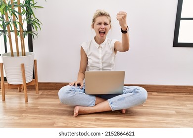 Young Blonde Woman Using Computer Laptop Sitting On The Floor At The Living Room Angry And Mad Raising Fist Frustrated And Furious While Shouting With Anger. Rage And Aggressive Concept. 