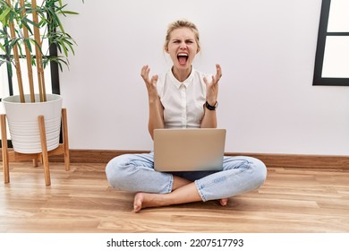 Young Blonde Woman Using Computer Laptop Sitting On The Floor At The Living Room Crazy And Mad Shouting And Yelling With Aggressive Expression And Arms Raised. Frustration Concept. 