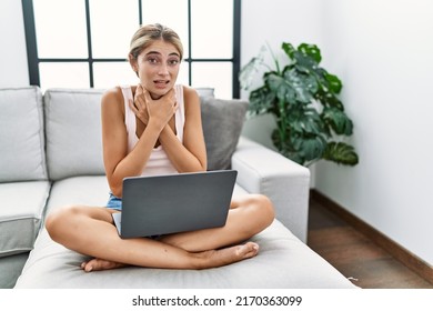Young blonde woman using laptop at home sitting on the sofa shouting and suffocate because painful strangle. health problem. asphyxiate and suicide concept. 