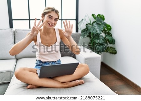 Young blonde woman using laptop at home sitting on the sofa showing and pointing up with fingers number eight while smiling confident and happy. 
