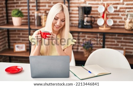 Young blonde woman using laptop drinking coffee sitting on table at home