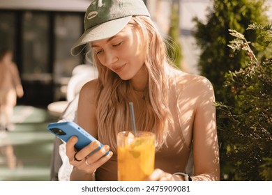 Young blonde woman using phone sits in cafe at table hold smartphone, answering texts, phone call, letters, posting photos, chatting or reading sms. Girl messaging, paying using phone, drink cocktail. - Powered by Shutterstock