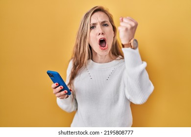 Young Blonde Woman Using Smartphone Typing Message Angry And Mad Raising Fist Frustrated And Furious While Shouting With Anger. Rage And Aggressive Concept. 