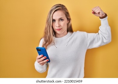 Young Blonde Woman Using Smartphone Typing Message Strong Person Showing Arm Muscle, Confident And Proud Of Power 