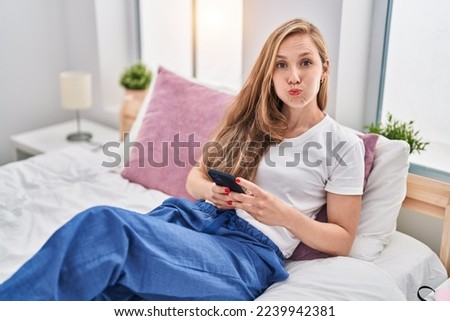 Young blonde woman using smartphone on bed puffing cheeks with funny face. mouth inflated with air, catching air. 