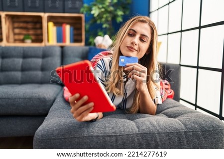 Young blonde woman using touchpad and credit card lying on sofa at home