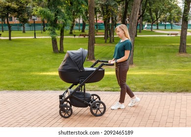 Young Blonde Woman Walking With Black Stroller In Summer Park. Happy Mother With Baby In Pram Outdoors. 