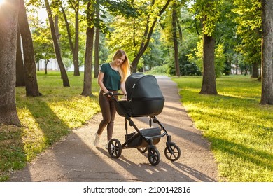 Young Blonde Woman Walking With Black Stroller In Summer Park. Happy Mother With Baby In Pram Outdoors. 