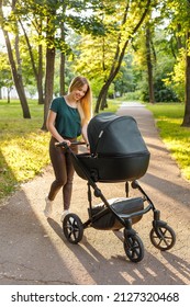 Young Blonde Woman Walking With Black Stroller In Summer Park. Happy Mother With Baby In Pram Outdoors. 