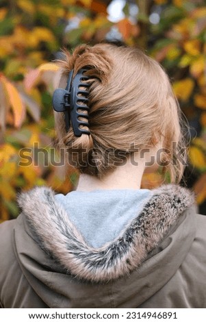 A young blonde woman walking outdoors with her back to camera showcasing black claw clip hairstyle. Casual hairstyle. Weekend hairstyle. Autumnal scene. Yellow and green leaves background. 