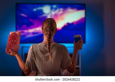 Young blonde woman watching TV in a cozy home environment with popcorn and a drink. Blue neon light. Evening. Watching TV programs, movies. Leisure at home, rest. - Shutterstock ID 2026428221