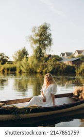 young blonde woman with wavy hair sits in a boat on the lake. girl in a long white dress posing in an old wooden boat. evening boat trip on the water near the house.