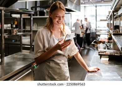 Young blonde woman wearing apron using cellphone while working in restaurant kitchen - Powered by Shutterstock