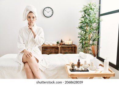 Young blonde woman wearing bathrobe at wellbeing spa asking to be quiet with finger on lips. silence and secret concept. 