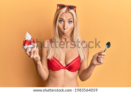 Young blonde woman wearing bikini holding ice cream afraid and shocked with surprise and amazed expression, fear and excited face. 