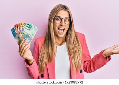 Young blonde woman wearing business style holding australian dollars celebrating achievement with happy smile and winner expression with raised hand 