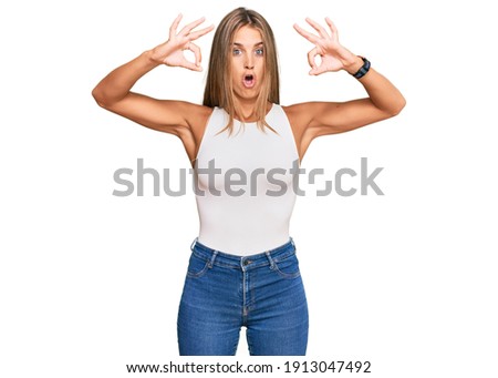 Young blonde woman wearing casual style with sleeveless shirt looking surprised and shocked doing ok approval symbol with fingers. crazy expression 
