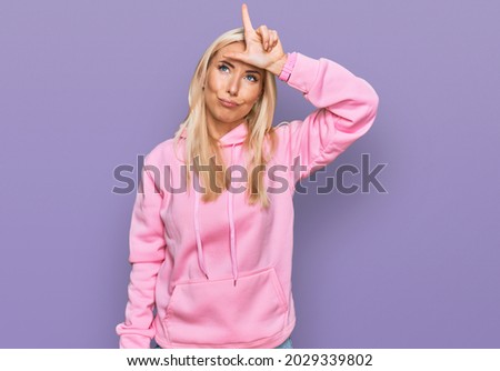 Young blonde woman wearing casual sweatshirt making fun of people with fingers on forehead doing loser gesture mocking and insulting. 