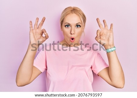 Young blonde woman wearing casual pink t shirt looking surprised and shocked doing ok approval symbol with fingers. crazy expression 