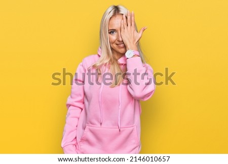 Young blonde woman wearing casual sweatshirt covering one eye with hand, confident smile on face and surprise emotion. 