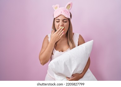 Young blonde woman wearing pyjama hugging pillow bored yawning tired covering mouth with hand. restless and sleepiness.  - Shutterstock ID 2205827799