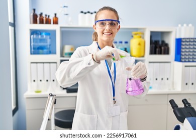 Young blonde woman wearing scientist uniform pouring liquid on test tube at laboratory - Shutterstock ID 2256917061