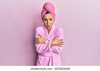 Young blonde woman wearing shower towel cap and bathrobe shaking and freezing for winter cold with sad and shock expression on face 