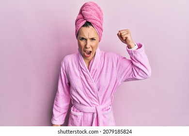 Young blonde woman wearing shower towel cap and bathrobe angry and mad raising fist frustrated and furious while shouting with anger. rage and aggressive concept. 