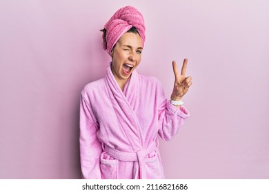 Young blonde woman wearing shower towel cap and bathrobe smiling with happy face winking at the camera doing victory sign with fingers. number two. 