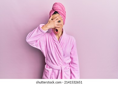 Young Blonde Woman Wearing Shower Towel Cap And Bathrobe Peeking In Shock Covering Face And Eyes With Hand, Looking Through Fingers With Embarrassed Expression. 