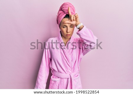 Young blonde woman wearing shower towel cap and bathrobe making fun of people with fingers on forehead doing loser gesture mocking and insulting. 