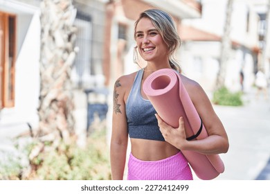 Young blonde woman wearing sportswear holding yoga mat at street - Powered by Shutterstock