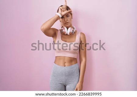 Young blonde woman wearing sportswear and headphones making fun of people with fingers on forehead doing loser gesture mocking and insulting. 