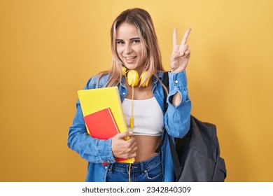 Young blonde woman wearing student backpack and holding books smiling looking to the camera showing fingers doing victory sign. number two. 