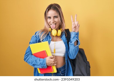 Young blonde woman wearing student backpack and holding books smiling with happy face winking at the camera doing victory sign with fingers. number two. 