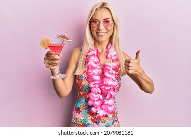 Young blonde woman wearing swimsuit and hawaiian lei drinking cocktail smiling happy and positive, thumb up doing excellent and approval sign 