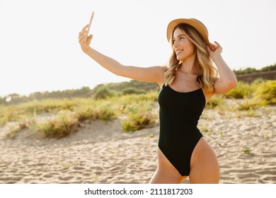 Young blonde woman wearing swimsuit taking selfie on cellphone at beach - Shutterstock ID 2111817203