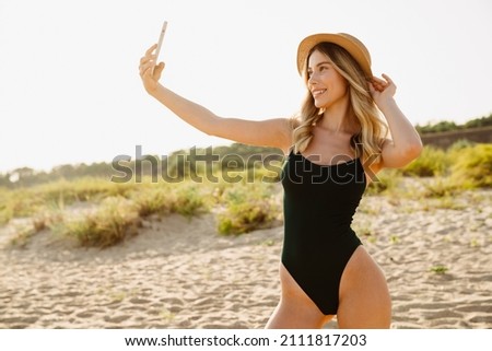 Young blonde woman wearing swimsuit taking selfie on cellphone at beach