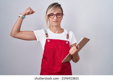 Young Blonde Woman Wearing Waiter Uniform Holding Clipboard Strong Person Showing Arm Muscle, Confident And Proud Of Power 