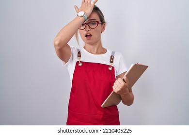 Young Blonde Woman Wearing Waiter Uniform Holding Clipboard Surprised With Hand On Head For Mistake, Remember Error. Forgot, Bad Memory Concept. 