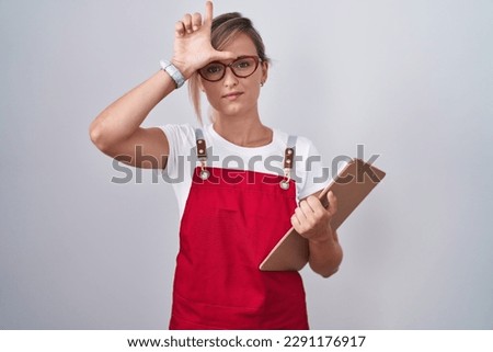Young blonde woman wearing waiter uniform holding clipboard making fun of people with fingers on forehead doing loser gesture mocking and insulting. 