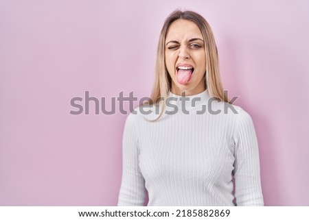 Young blonde woman wearing white sweater over pink background sticking tongue out happy with funny expression. emotion concept. 