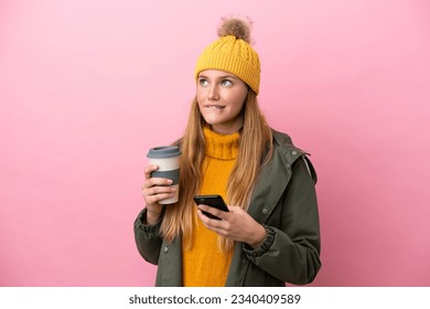 Young blonde woman wearing winter jacket isolated on pink background holding coffee to take away and a mobile while thinking something - Powered by Shutterstock