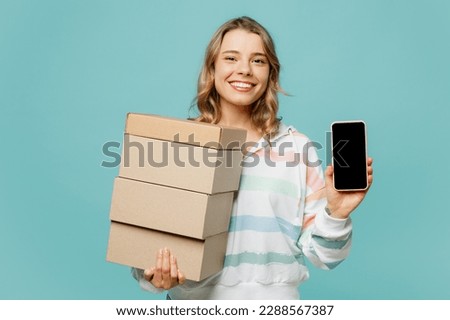 Young blonde woman wears striped hoody use mobile cell phone with blank screen workspace area hold stack cardboard blank boxes isolated on plain pastel light blue cyan background. Lifestyle concept