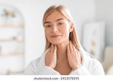 Young blonde woman in white bathrobe massaging skincare lotion on her neck, moisturizing skin and promoting daily wellness and beauty routine in modern bathroom interior, looking at camera