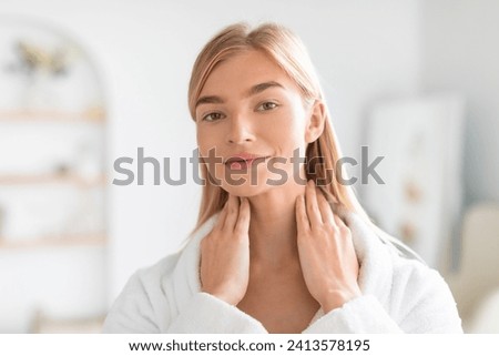Young blonde woman in white bathrobe massaging skincare lotion on her neck, moisturizing skin and promoting daily wellness and beauty routine in modern bathroom interior, looking at camera