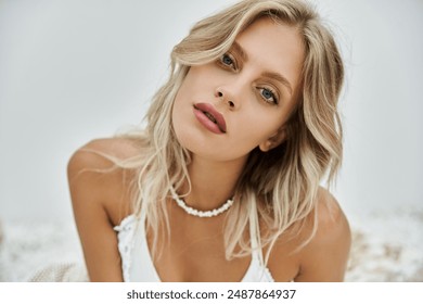 A young blonde woman in a white beach top poses against a grey backdrop, showcasing summer fashion and trends.