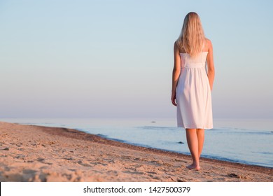 Young blonde woman in white dress walking alone on sand beach. Summer evening. Fresh air. Orange sunset light. Back view. Empty place for emotional, sentimental, inspirational text, quote or sayings.